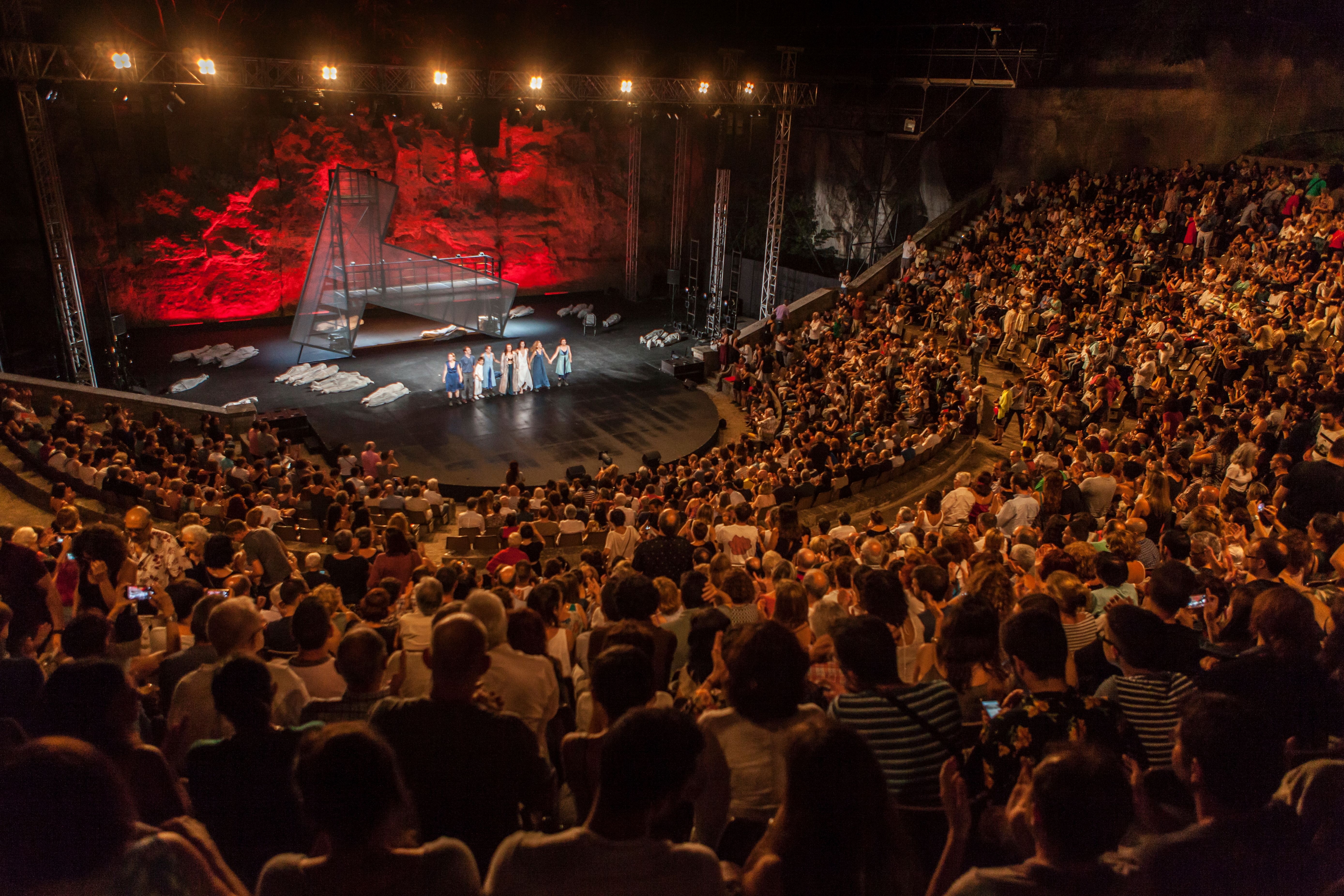 Gran teatre a l’aire lliure ple de públic assegut en grades semicirculars. A l’escenari central, diverses actrius saluden mentre al terra hi ha figures estirades cobertes amb teles clares. El fons de l’escenari està il·luminat amb llums vermelles intenses sobre una paret rocosa. 