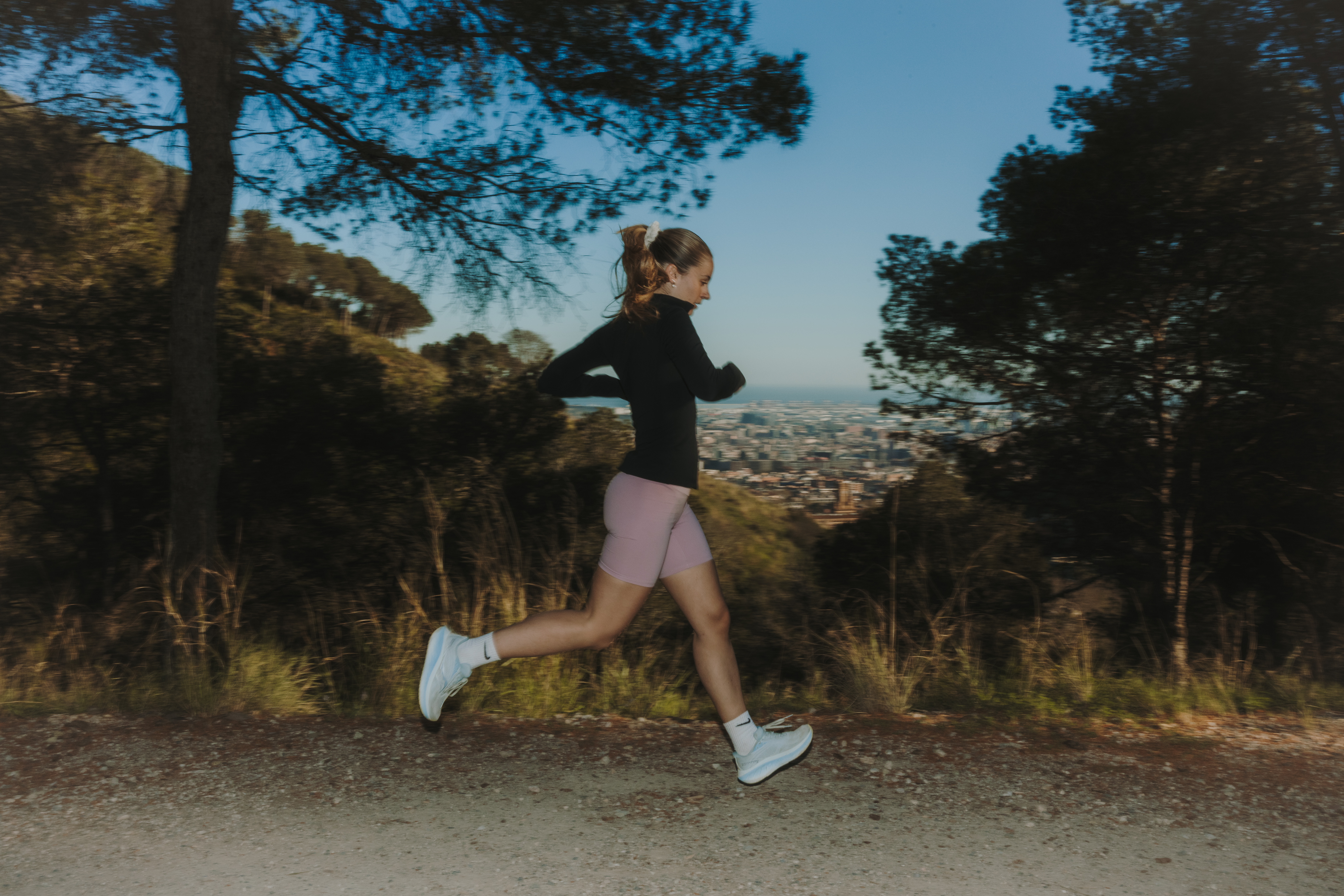 Chica corriendo por un camino de tierra entre árboles, con una vista panorámica de Barcelona al fondo.