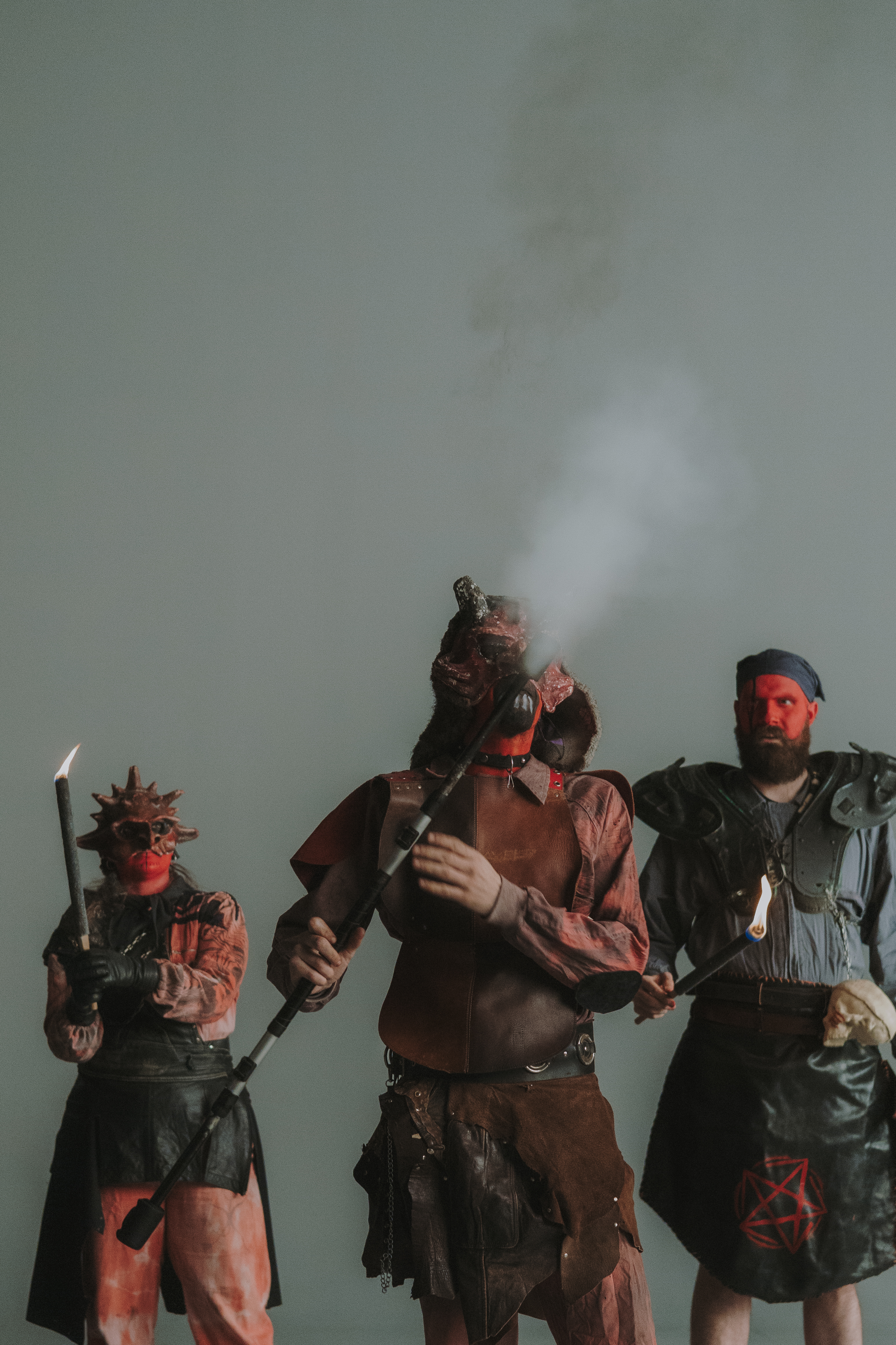 Trois personnes déguisées en diables tiennent des torches et un bâton fumant, avec des visages peints ou masqués et des vêtements de protection. La scène, sur un fond neutre, évoque la tradition du correfoc et l’univers festif du feu.
