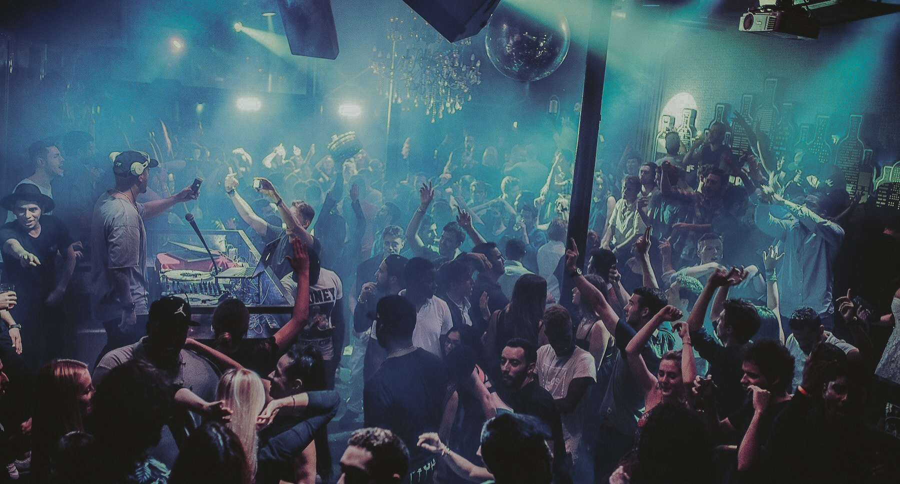 Discoteca plena de gent ballant sota llums blaves i fum, amb un DJ actuant en una cabina elevada mentre el públic aixeca les mans o grava amb el mòbil, creant un ambient festiu i vibrant de nit.