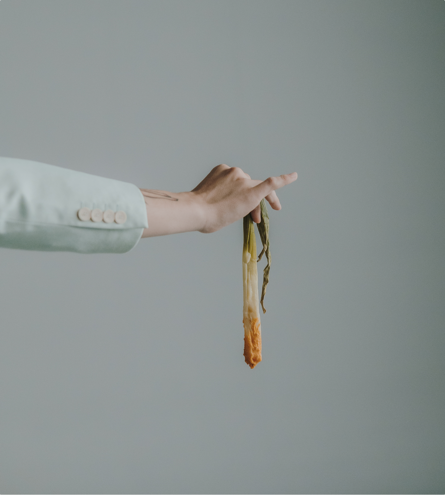 El braç d'una persona que porta una jaqueta amb les mànigues de color clar aguanta un calçot sucat en salsa. El fons és llis i neutre, de color gris clar.