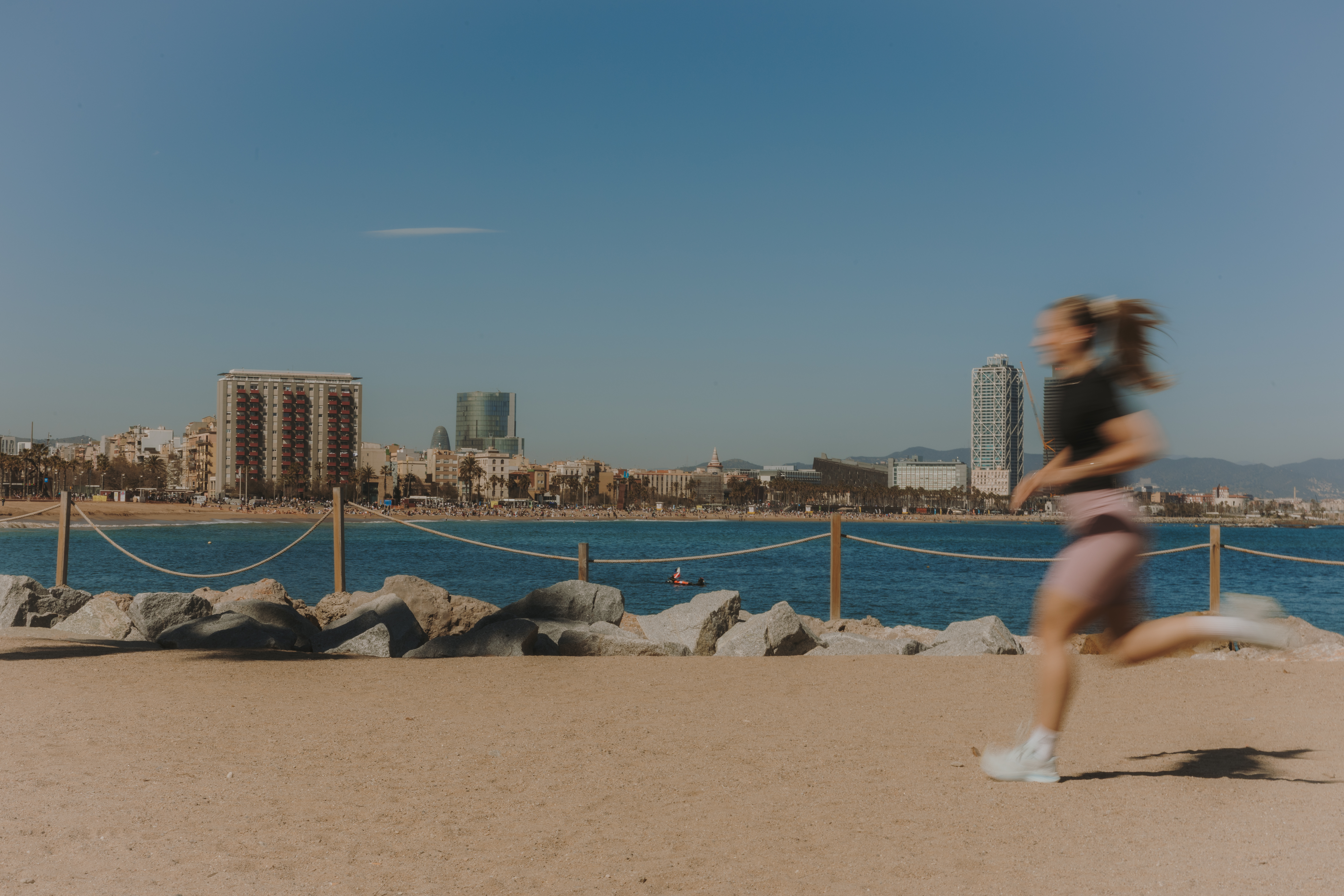 Noia corrent pel passeig a costat del mar. De fons, diversos edificis de l'skyline de Barcelona sota un cel blau sense núvols en un dia de sol.