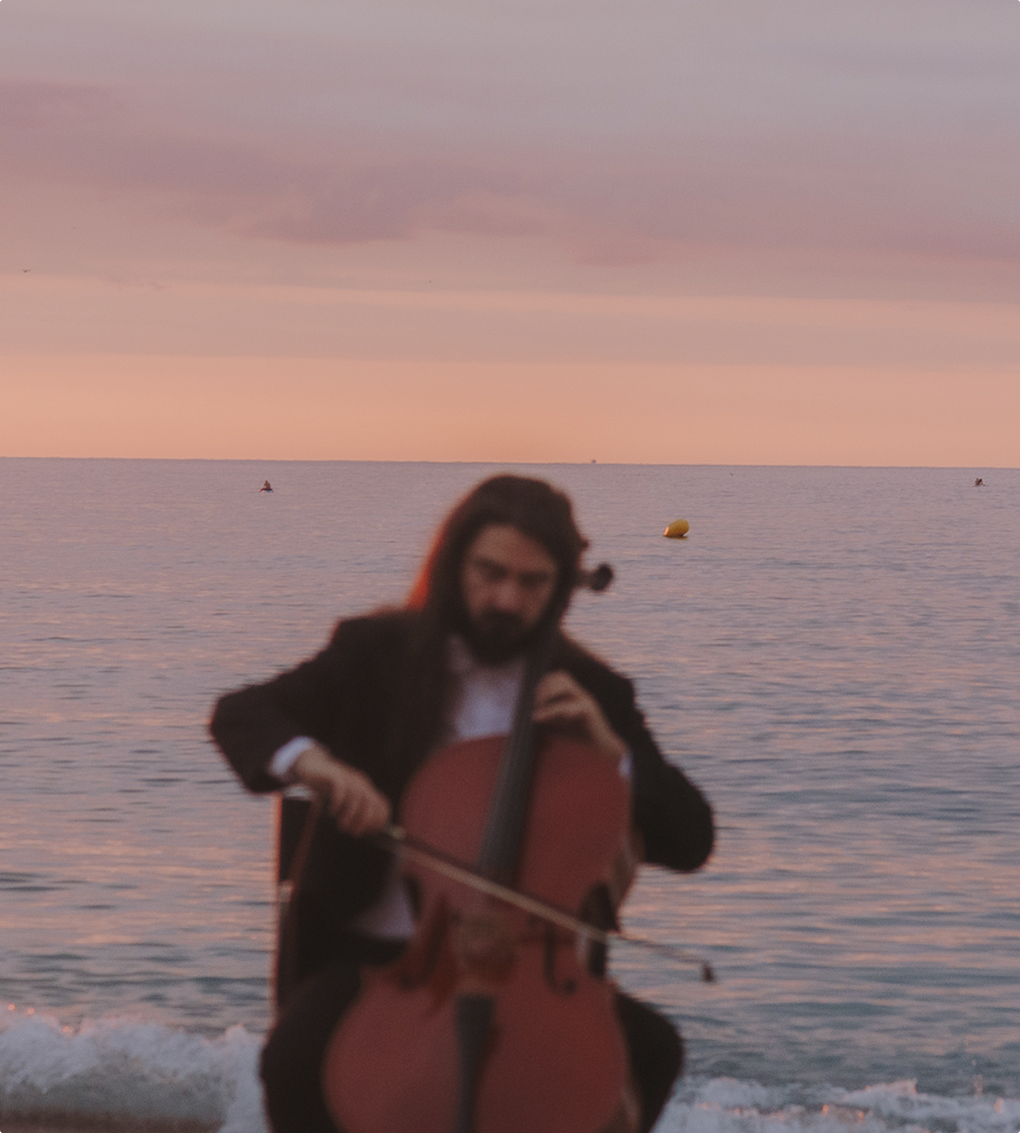 Un home amb barba i vestit formal toca el violoncel a la platja, amb el mar al fons i un cel de capvespre.