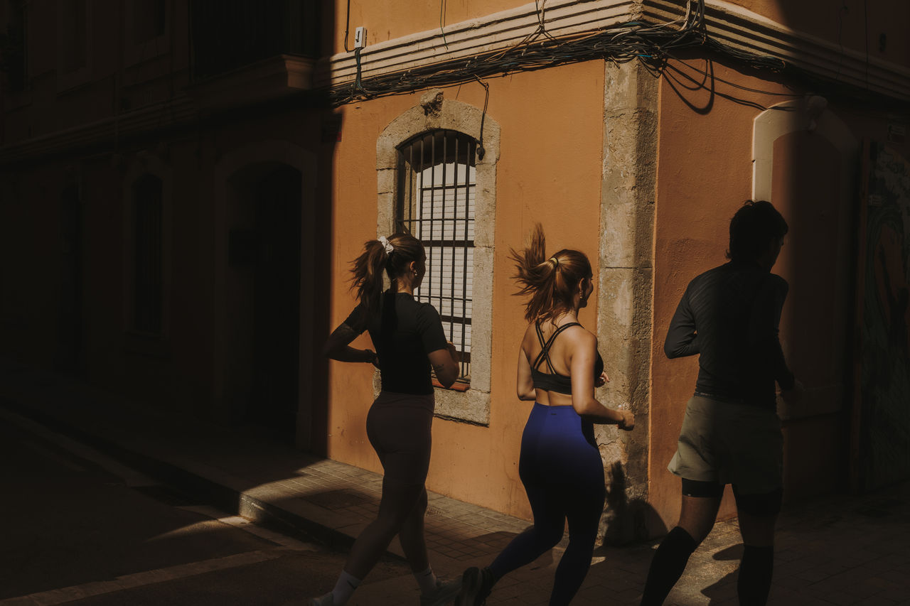 Tres persones corren per un carrer de Barcelona il·luminat pel sol, al costat de la façana d’un edifici.