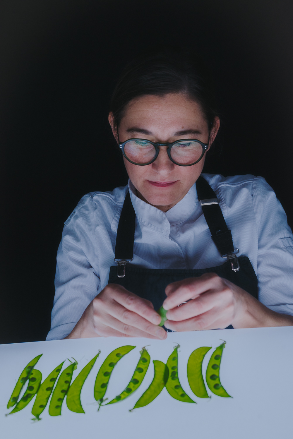 Xef amb ulleres i uniforme de cuina manipulant pèsols verds sobre una taula il·luminada, amb fons negre.