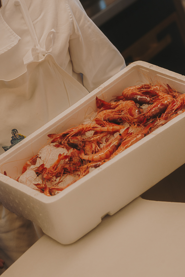 Persona sostenint una caixa blanca de porexpan amb diverses gambes vermelles fresques disposades sobre gel. La imatge mostra un primer pla del recipient en un entorn de preparació gastronòmica.