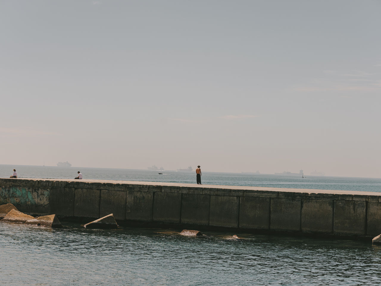 Persones passejant i assegudes sobre un espigó davant del mar de Barcelona, amb vaixells a l’horitzó i aigües tranquil·les en primer pla.
