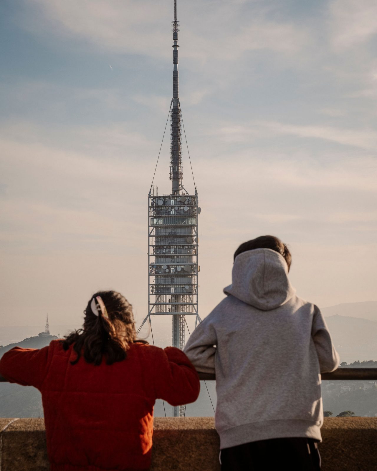Dues persones d’esquena observant la torre de comunicacions de Collserola des d’un mirador de Barcelona, amb el paisatge urbà i les muntanyes al fons.