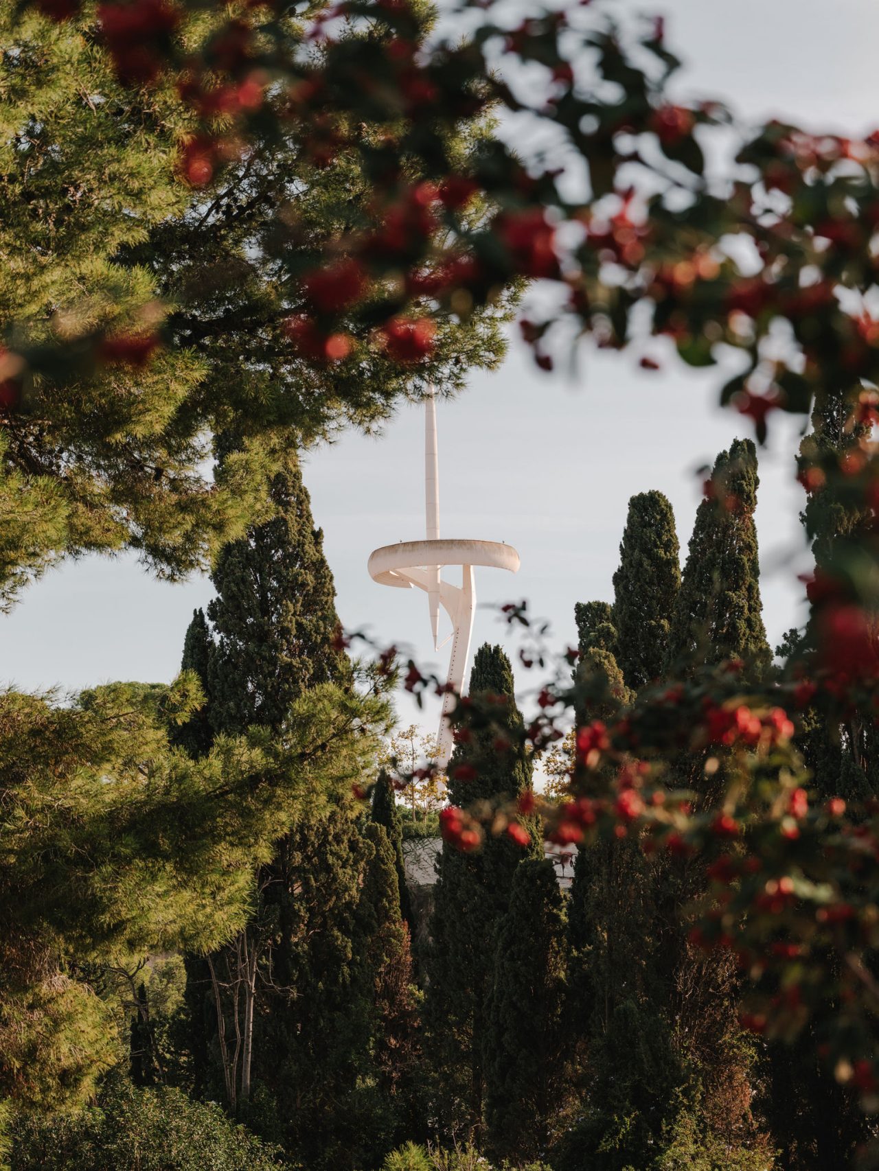 Torre de comunicacions de Montjuïc envoltada de vegetació, vista entre arbres i flors vermelles desenfocades en primer pla.
