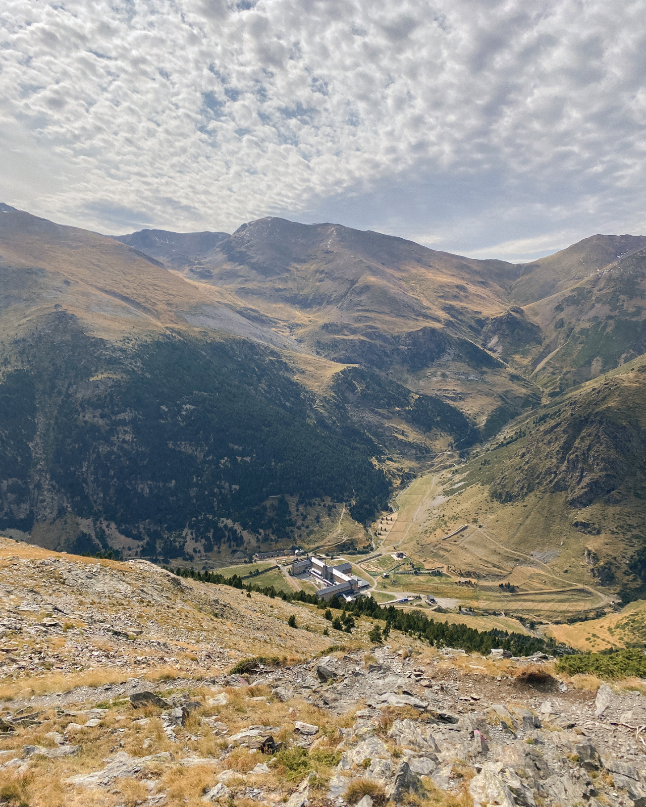 Vista panoràmica d’una vall de muntanya amb camins, edificis i relleu ondulat sota un cel ennuvolat.