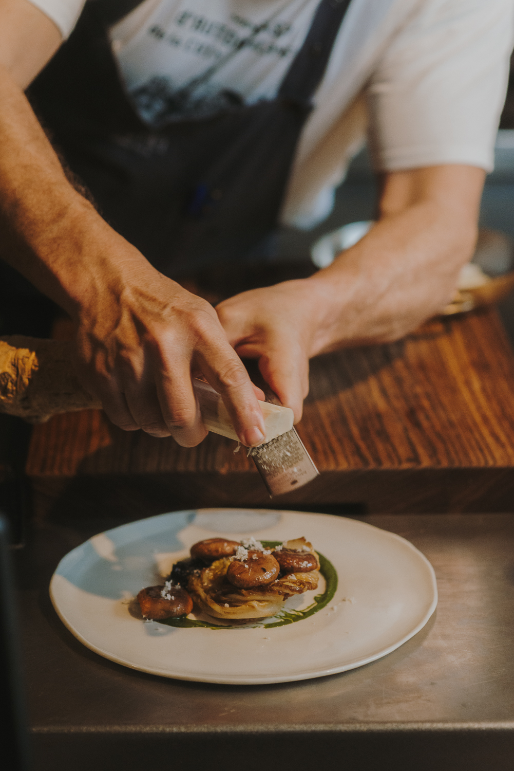 Chef’s hands decorating the dish by grating cheese. Between the chef and the plate is a wooden cutting board.