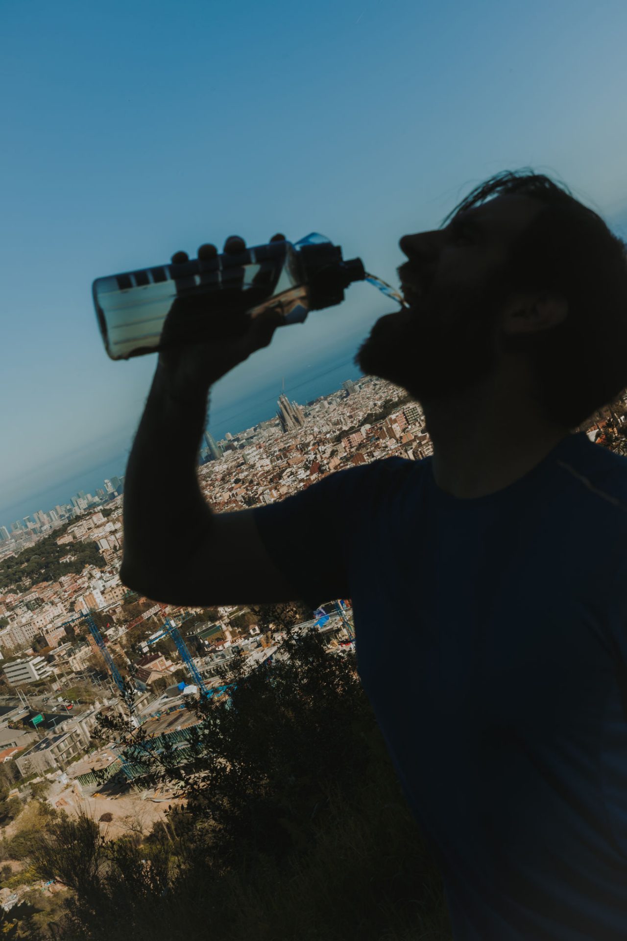 Man drinking water from a bottle in an elevated area, with a panoramic view of Barcelona and the sea in the background.