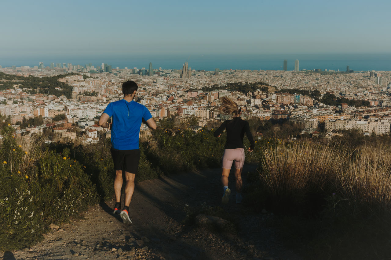 Two people running along a dirt path with a panoramic view of Barcelona, the Sagrada Família and the sea in the background.
