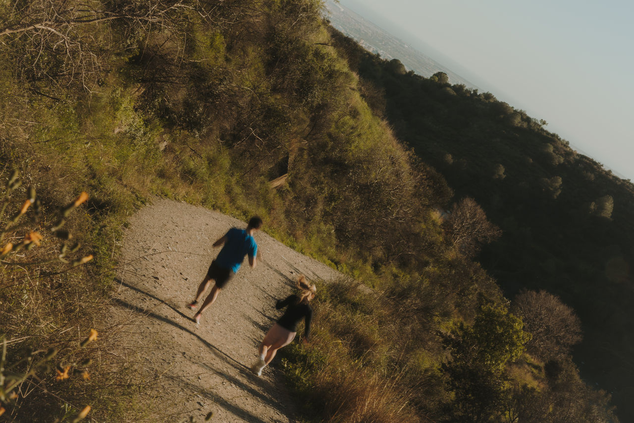 Two people running along a mountain path surrounded by vegetation, with the Collserola hillside in the background.
