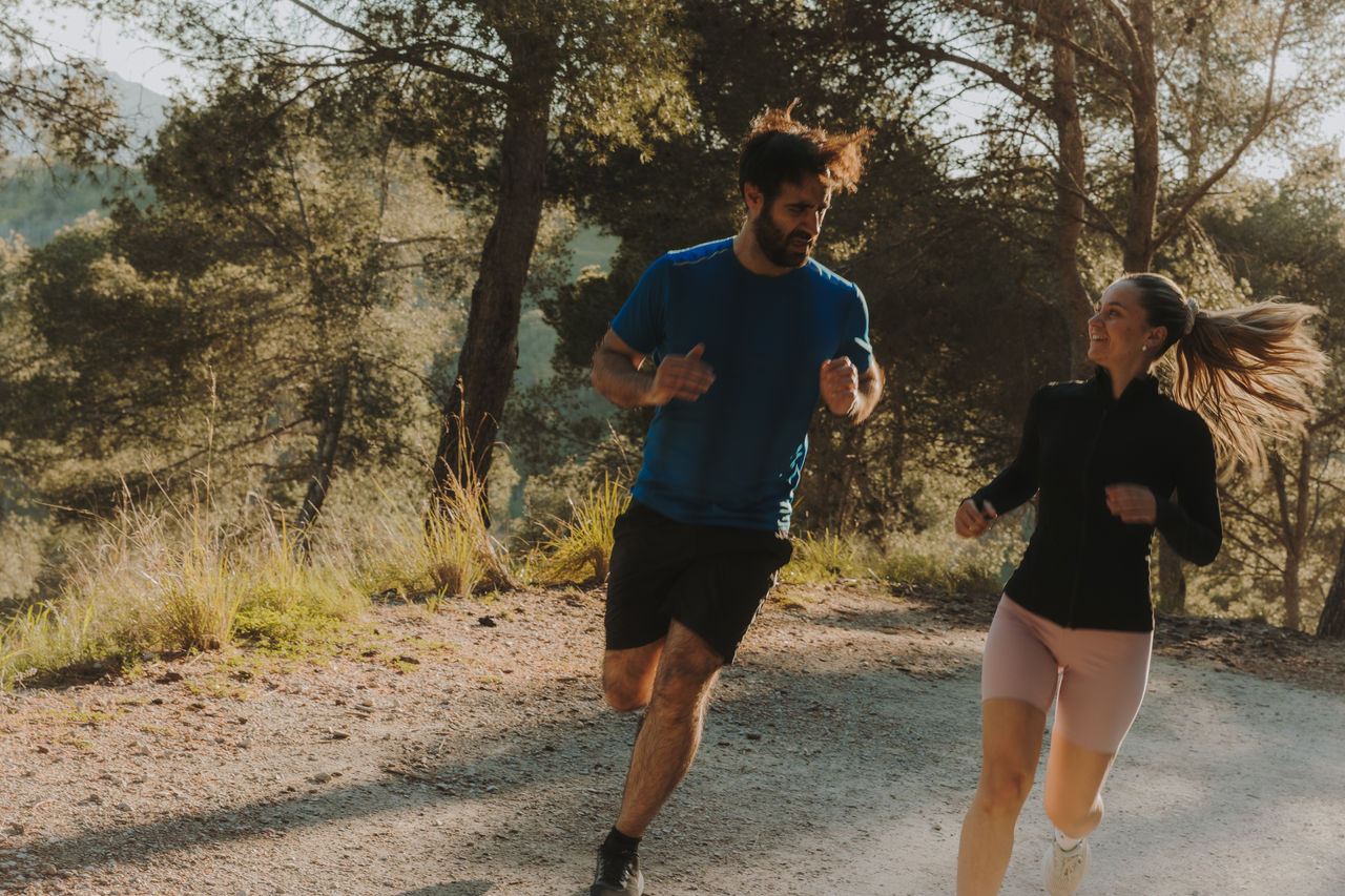 Two people running along a dirt path in a wooded area, surrounded by trees and vegetation.
