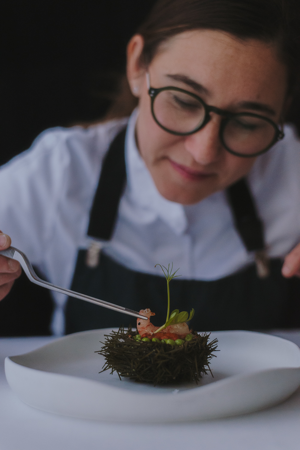 Chef con gafas decorando un plato gastronómico con guisantes y brotes verdes sobre una base oscura, en una presentación cuidada.
