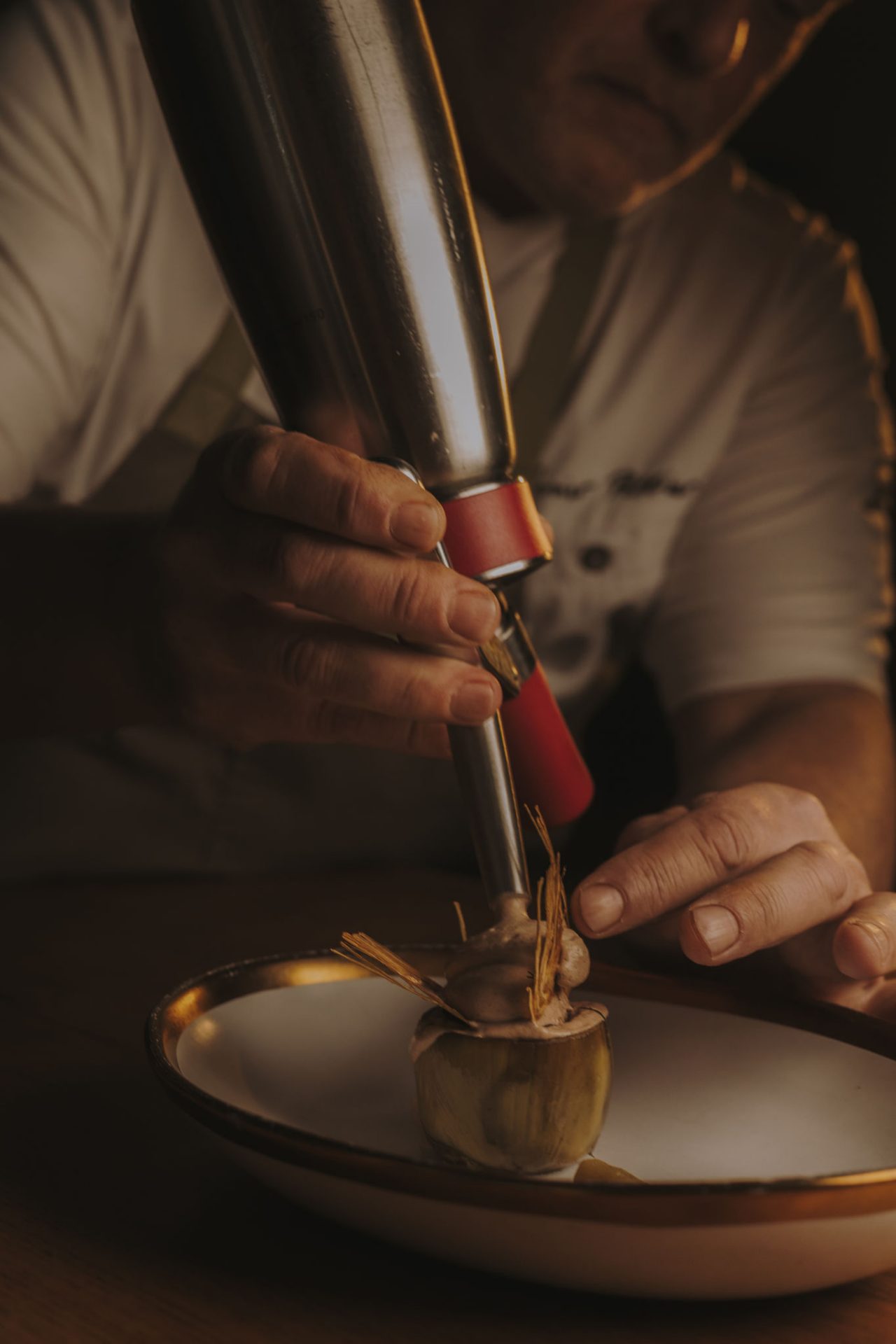 Manos de un chef usando un sifón culinario para terminar un plato de alcachofa en un plato decorativo. El chef lleva una camiseta de manga corta y un delantal gris. La luz es cálida.