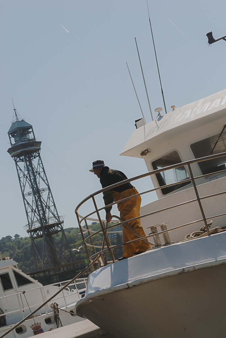 Hombre trabajando en la cubierta de una embarcación en el puerto de Barcelona, con la torre del teleférico del puerto al fondo. Lleva ropa de trabajo naranja y negra y está apoyado en la barandilla del barco en un día soleado.