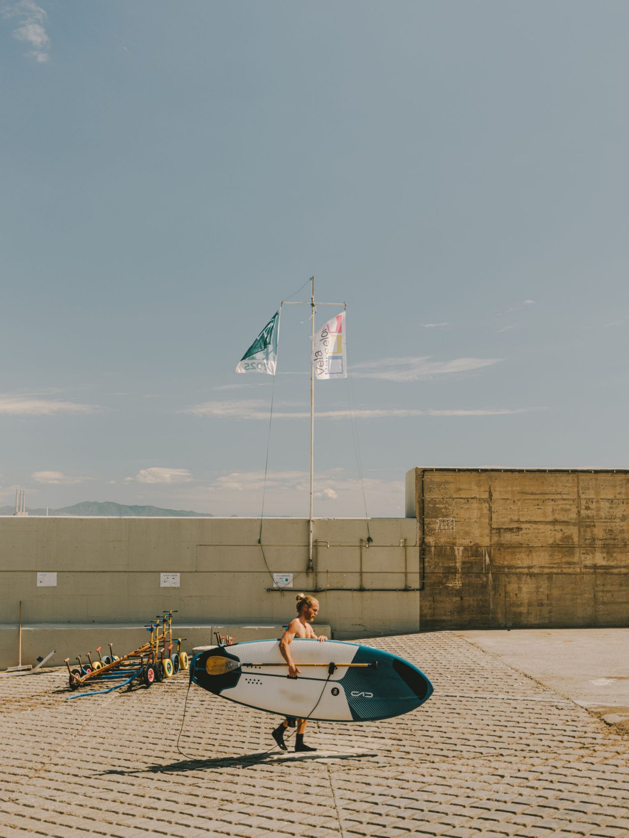 Personne portant une planche de paddle dans une zone pavée près de la mer, avec des drapeaux, des pagaies et l’horizon maritime en arrière-plan.