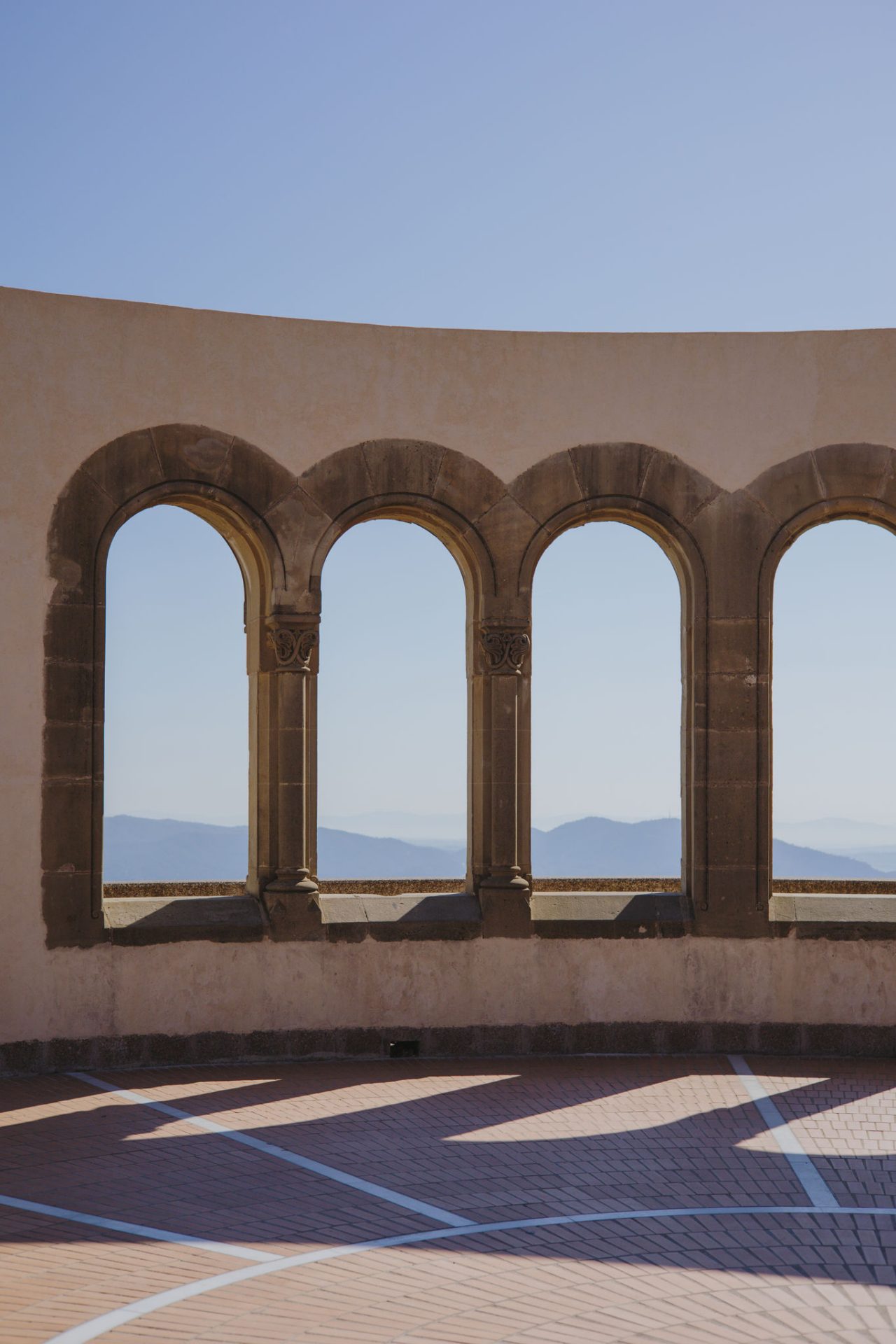 Arcades en pierre avec vue sur les montagnes depuis un belvédère, avec une lumière nette et des ombres marquées au sol.