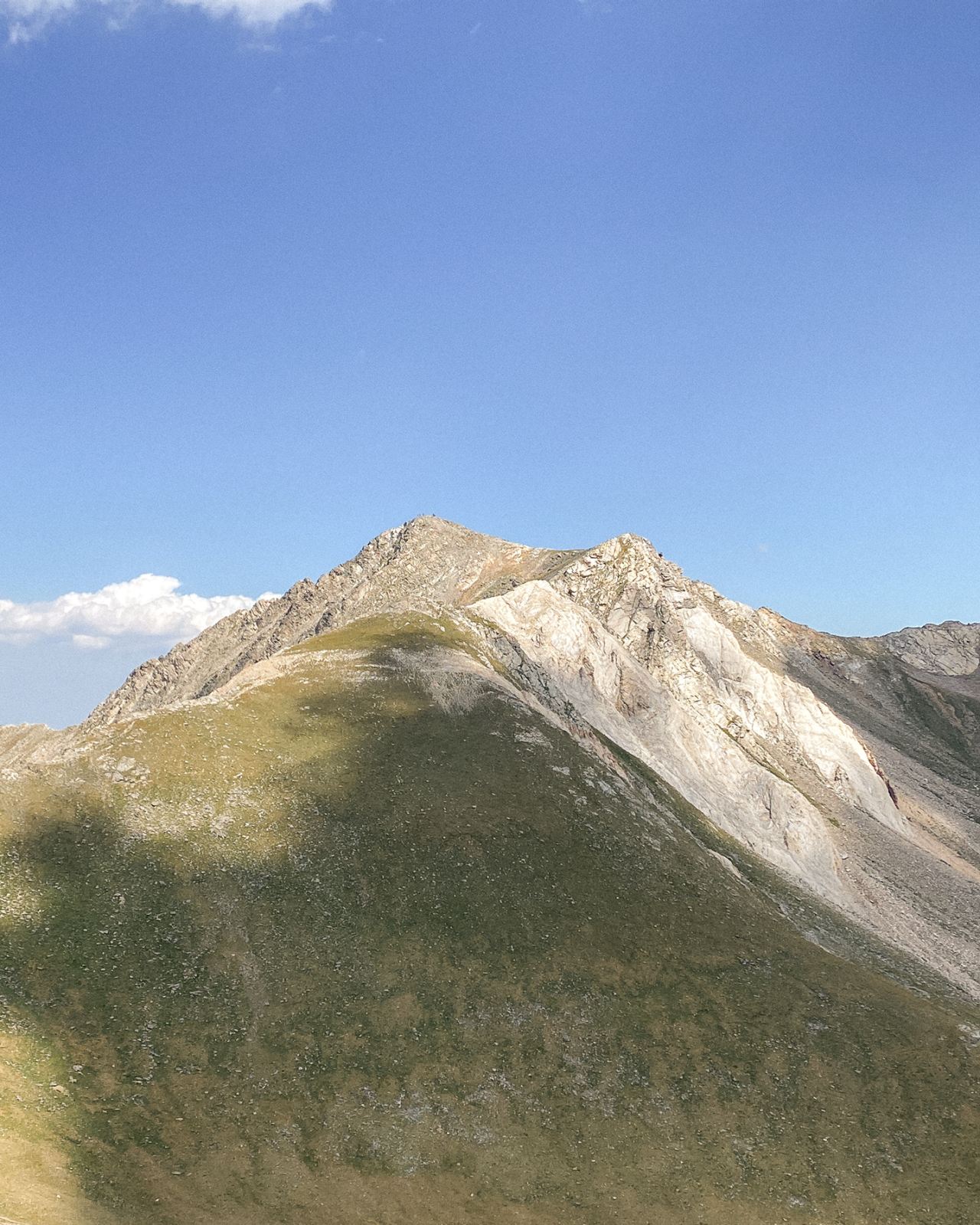 Sommet de montagne avec des pentes herbeuses et rocheuses sous un ciel bleu avec des nuages dispersés