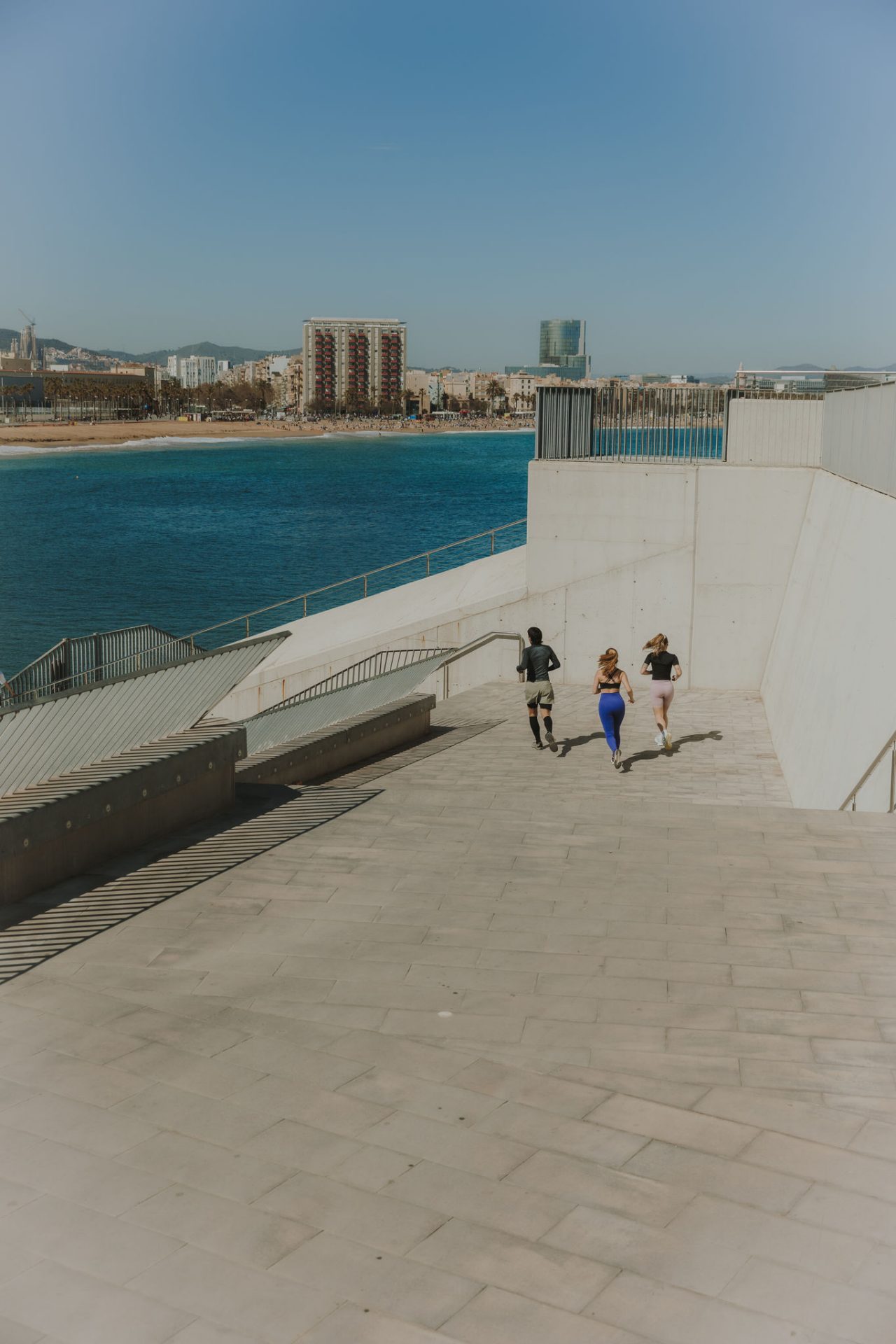 Trois personnes courent dans des escaliers au bord de la mer, avec la plage et les bâtiments de Barcelone à l’arrière-plan.