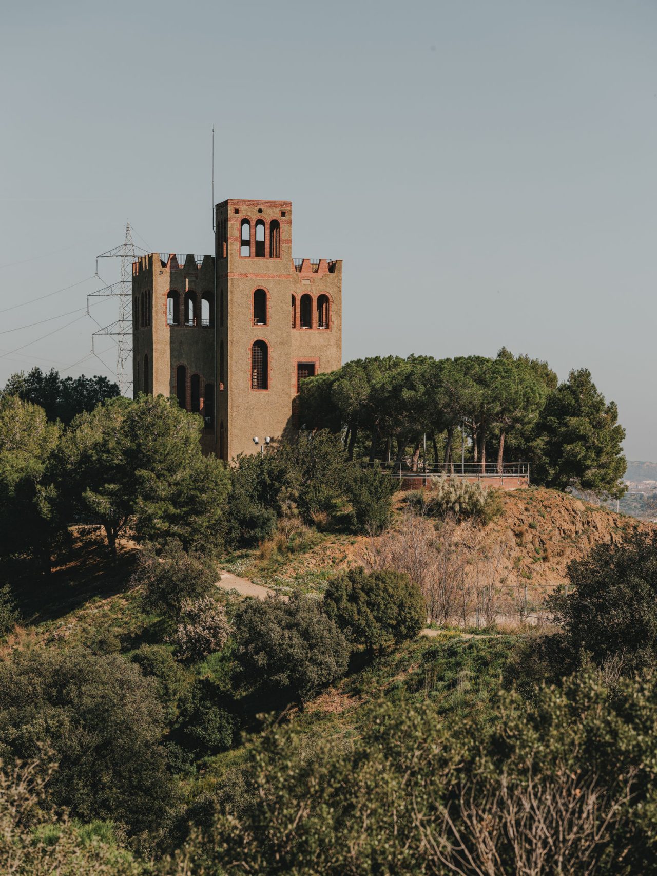 Torre di avvistamento in mattoni situata su una collina di Collserola, circondata da pini e vegetazione mediterranea, con una linea elettrica sullo sfondo