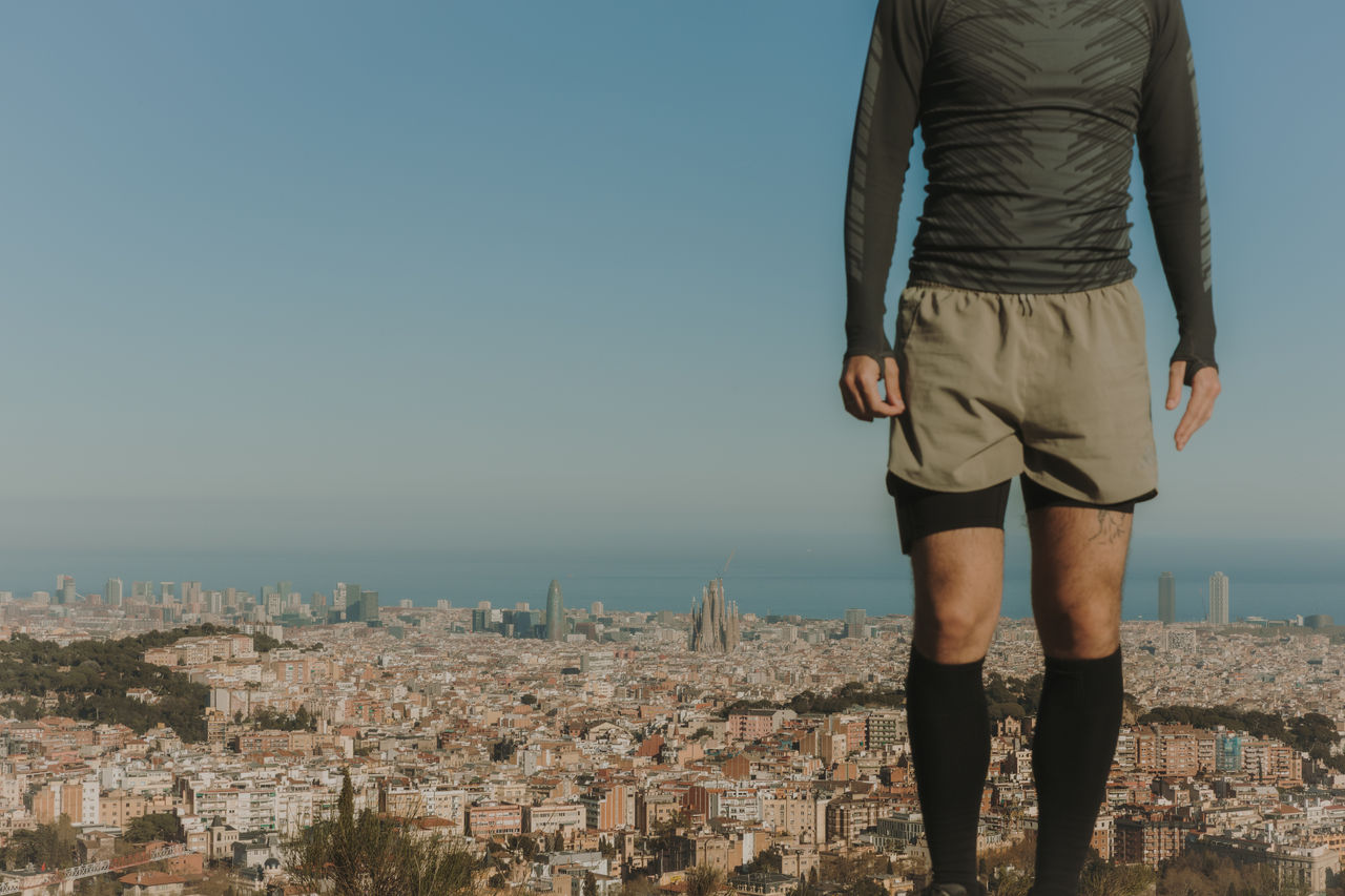 Uomo in piedi in una zona elevata di montagna, con una vista panoramica di Barcellona, la Sagrada Família e il mare sullo sfondo.